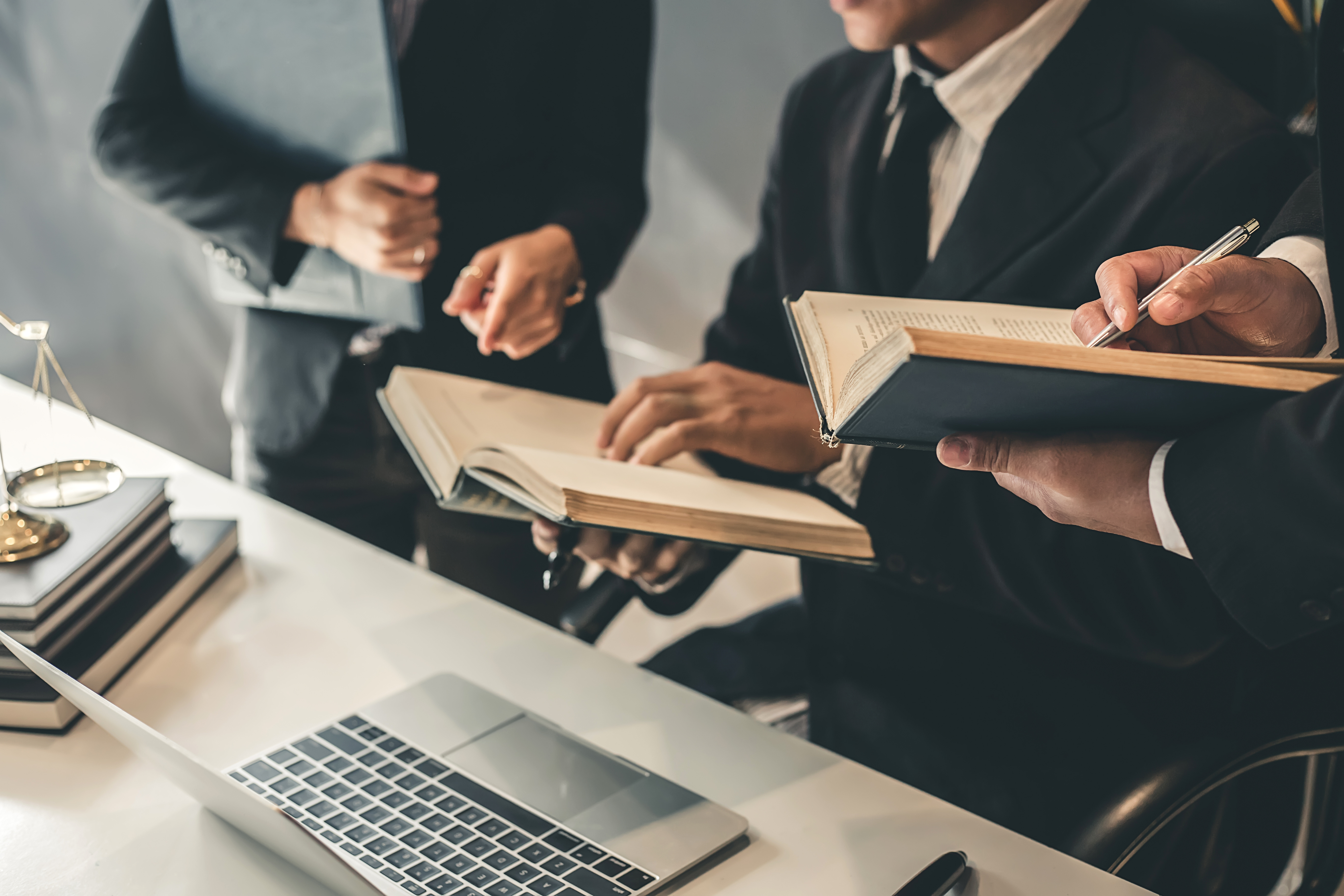Group of lawyers or legal professionals reviewing open law books and taking notes at a desk with a laptop, scales of justice, and documents, representing teamwork and legal research.